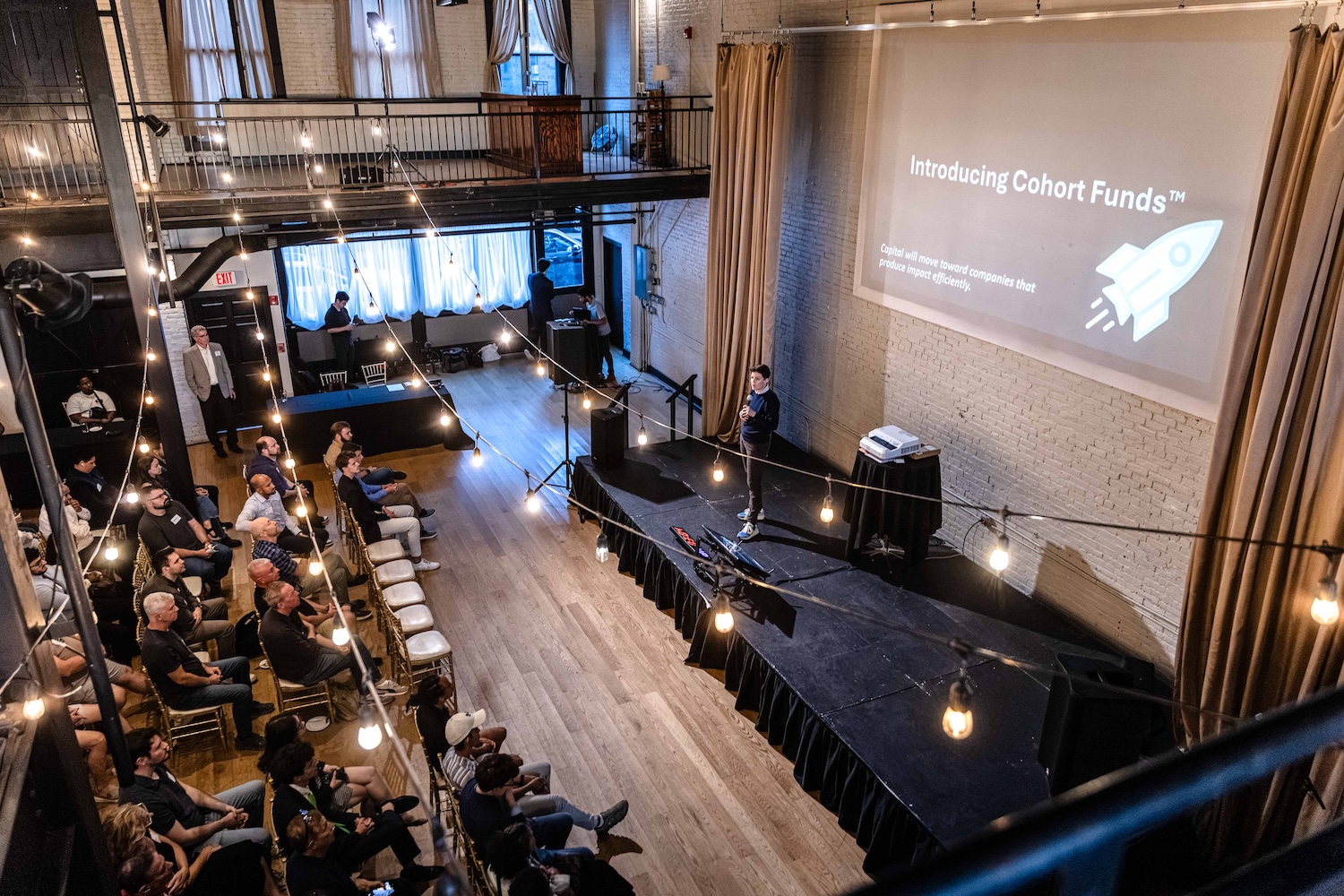 A speaker presents “Introducing Cohort Funds” to an audience in a loft-style venue with string lights and a projection screen.