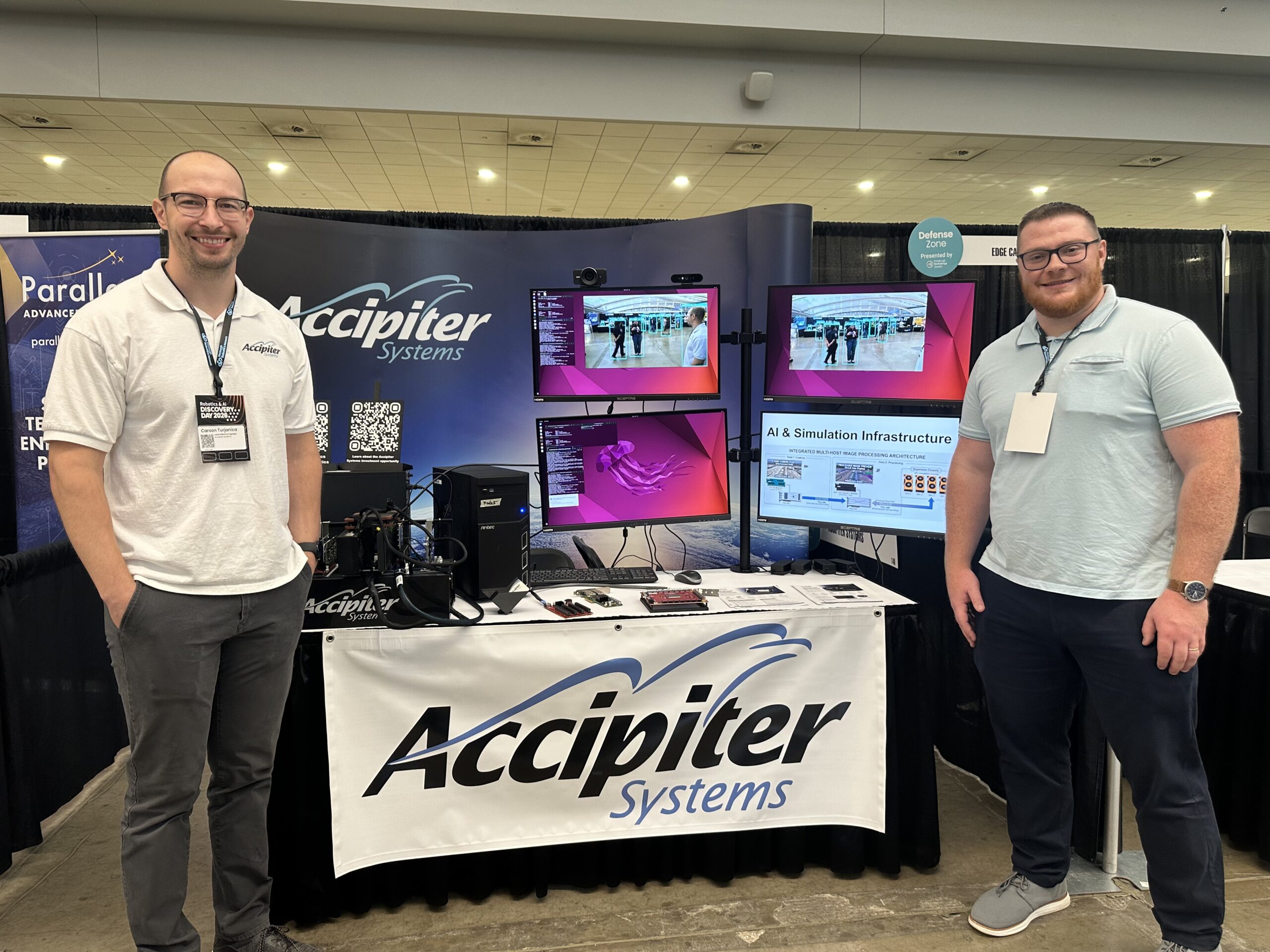 Two men stand at an Accipiter Systems booth featuring computer monitors, equipment, and informational displays at a convention or trade show.