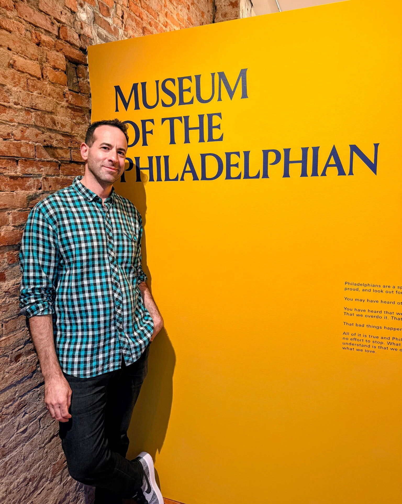 A man in a green plaid shirt leans against a yellow wall with the text “Museum of the Philadelphian” printed on it.
