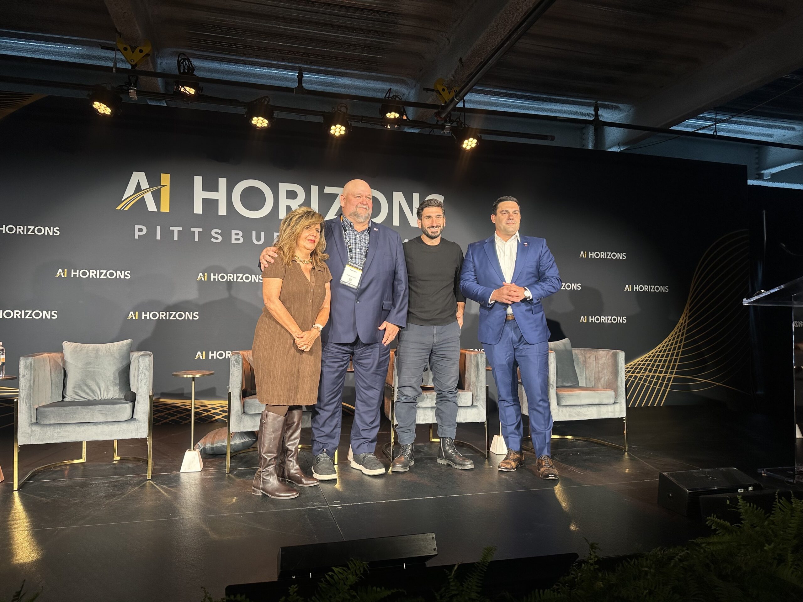 Four people stand on stage in front of an “AI Horizons Pittsburgh” backdrop, facing the camera, with chairs and stage lights visible behind them.