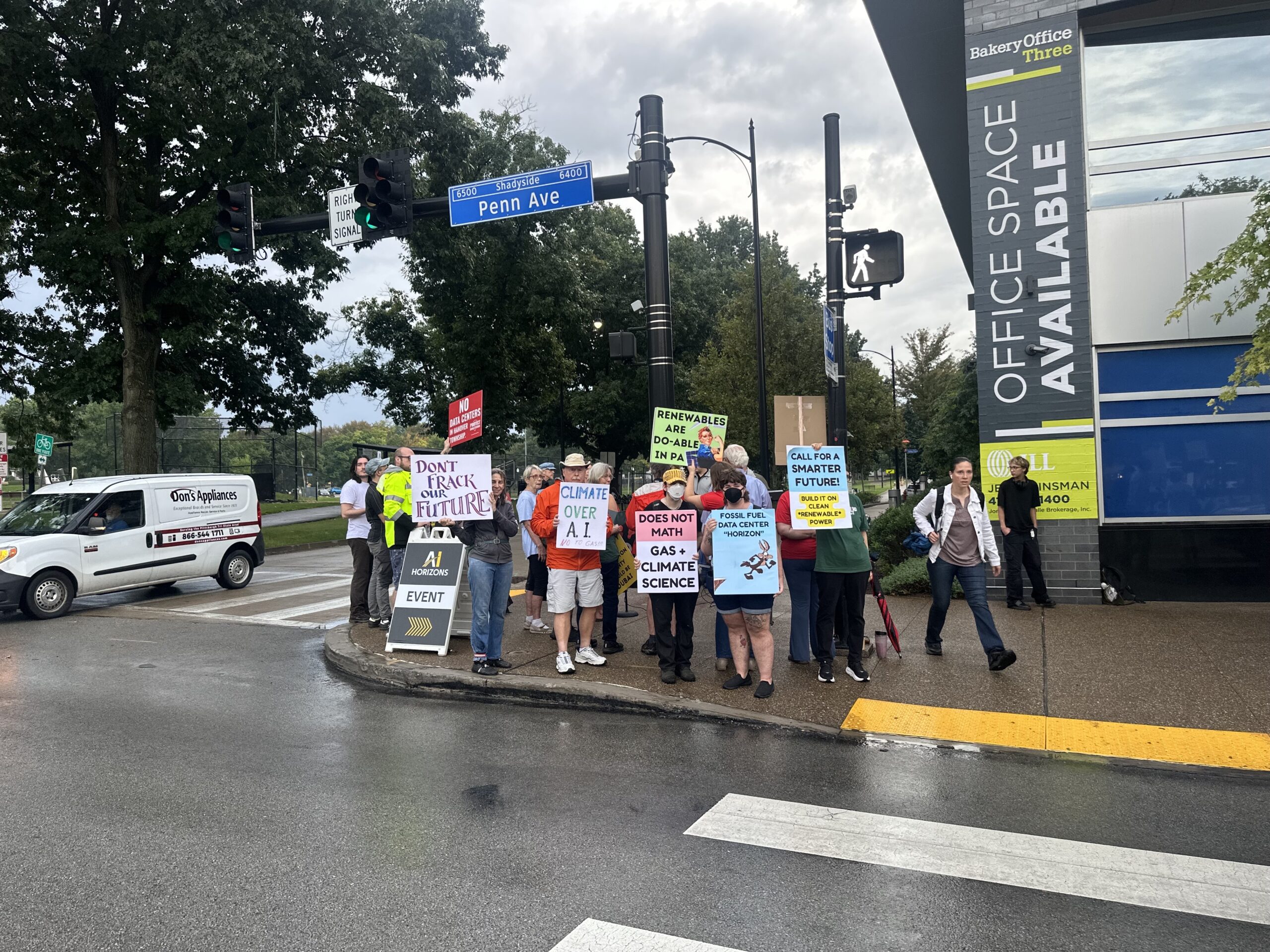 A group of people holding protest signs about climate change and AI stand on a city street corner near a Penn Ave sign and an office space building.