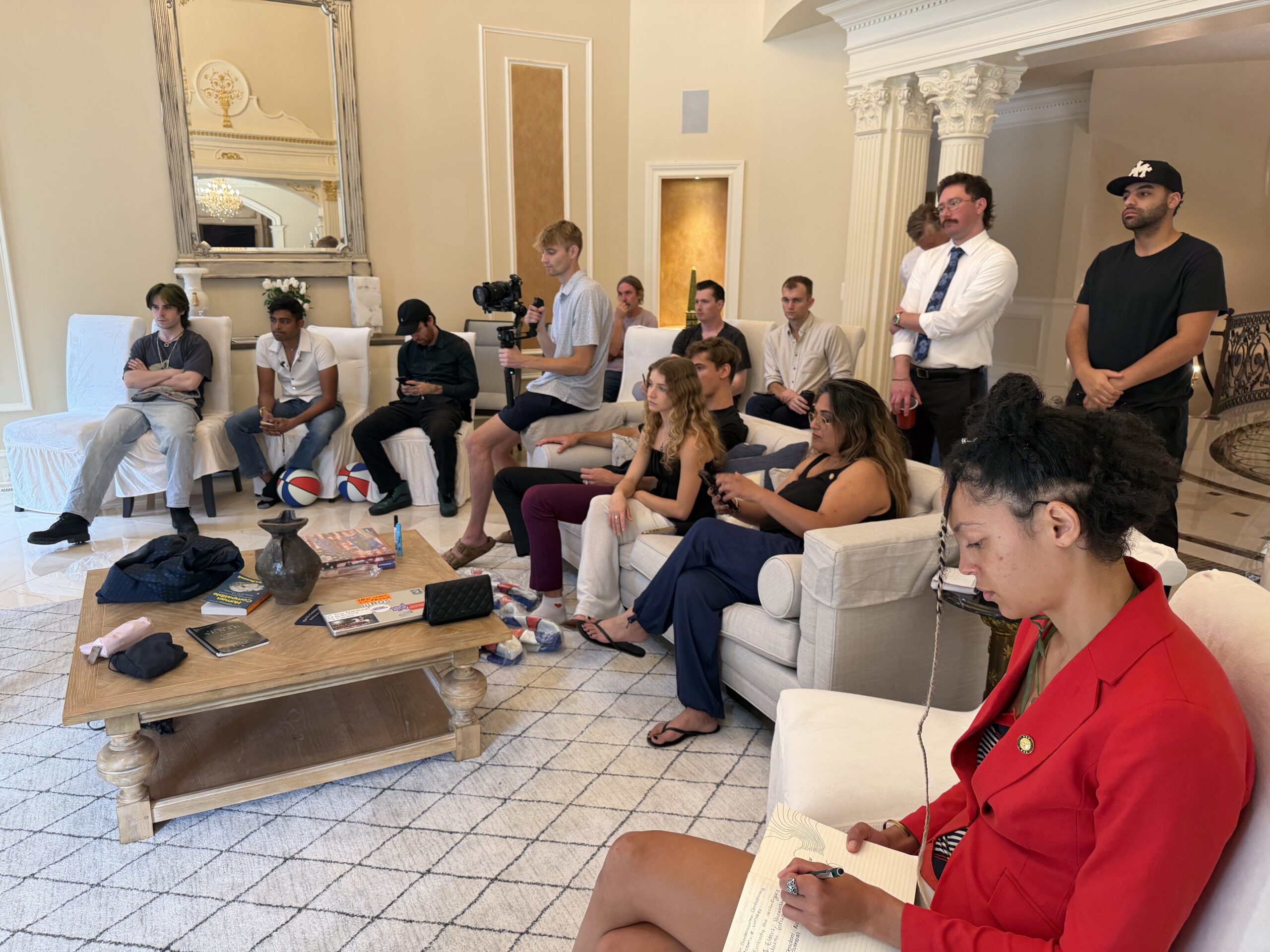 A group of people sits and stands in a well-lit living room, some taking notes, others watching attentively, with cameras and notebooks visible.