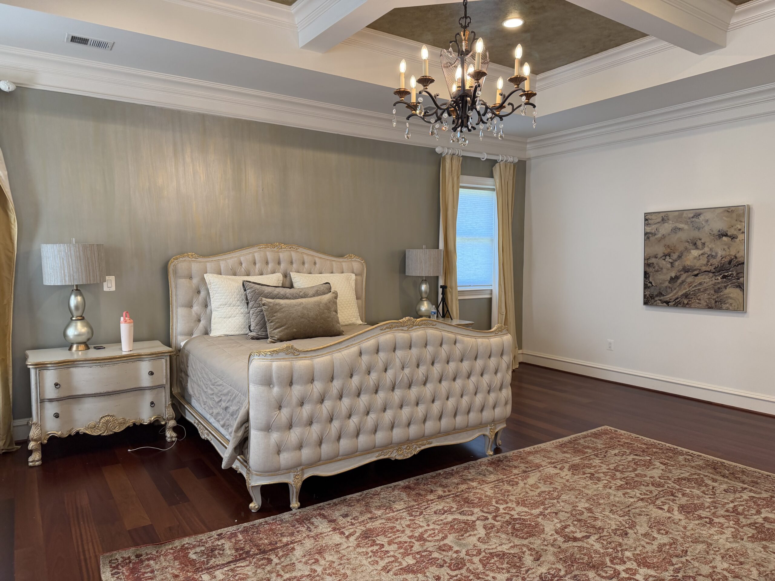 Elegant bedroom with a tufted bed, two lamps on nightstands, a chandelier, large rug, curtains, and a wall painting. A red-and-white bottle sits on the left nightstand.