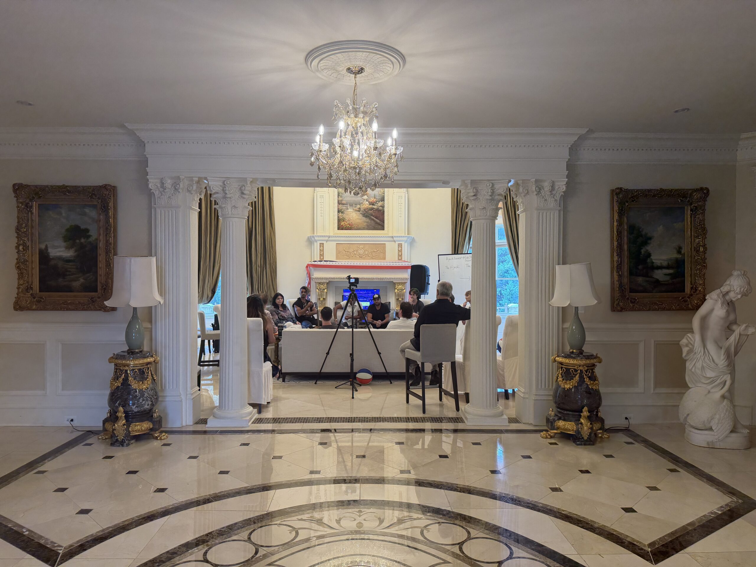 A group of people sit at tables in an ornate room with columns, paintings, chandeliers, a statue, and a camera set up, engaged in a meeting or presentation.