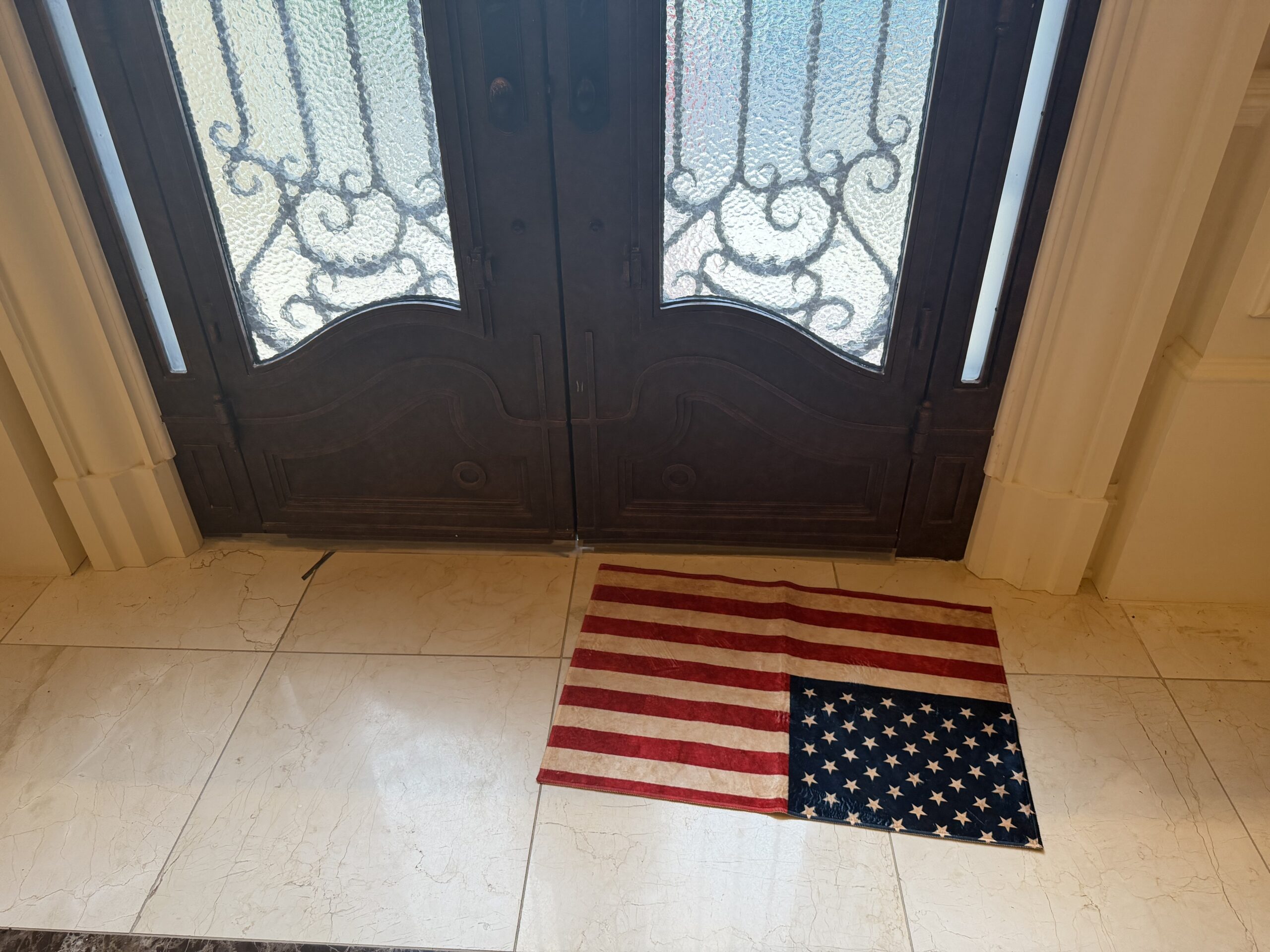 An American flag is laid flat on a tiled floor in front of double wooden doors with glass panels.
