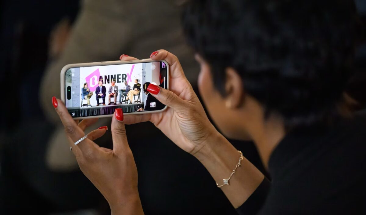 A person with red nail polish holds a smartphone, recording or photographing a panel discussion with four speakers on stage.