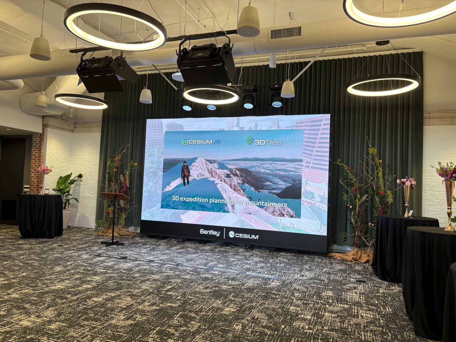 A conference room with a large screen displaying a mountain climber and the text “3D expedition planning for mountaineers” beneath company logos.