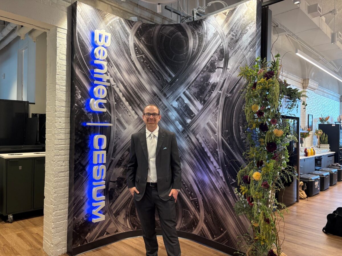 A man in a suit stands in front of a Bentley | Cesium backdrop with an abstract design, next to a vertical floral arrangement indoors.