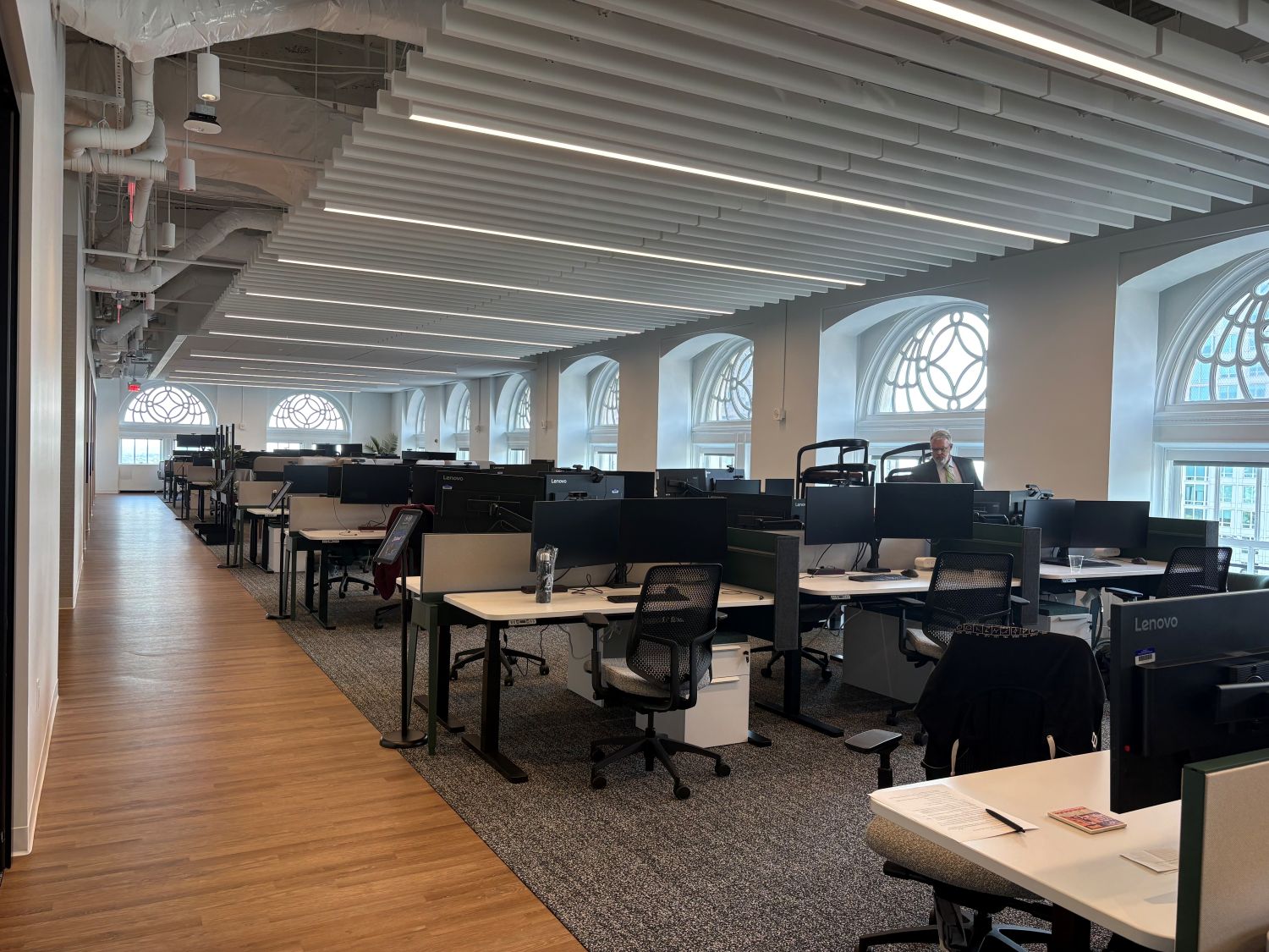 Modern open-plan office with rows of desks, computer monitors, and chairs. Large arched windows let in natural light. One person is standing near the back of the room.