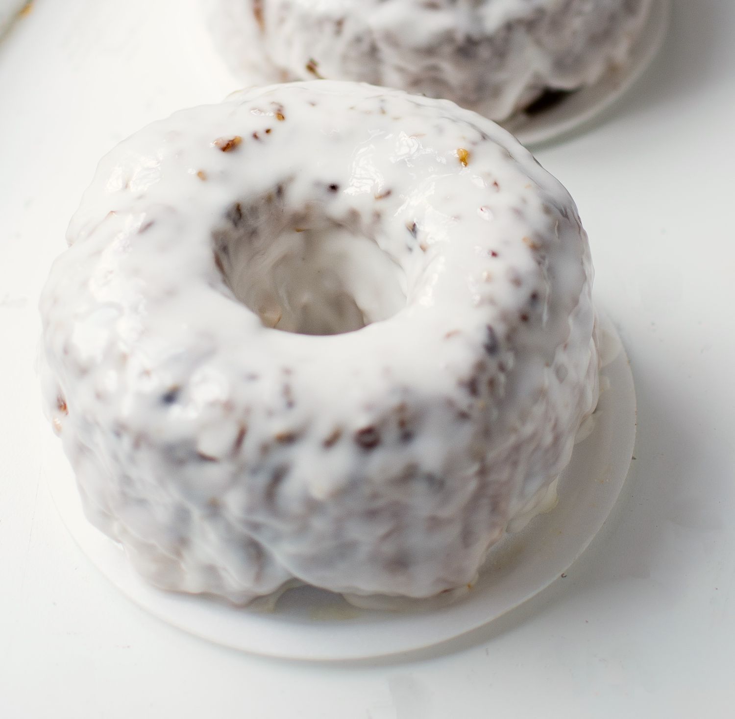 Small bundt cake covered in a thick white glaze, placed on a white surface with part of another cake visible in the background.