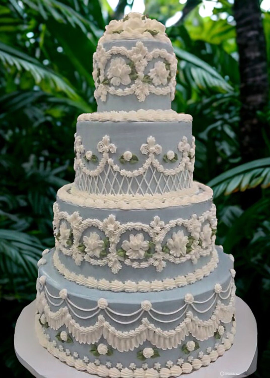 Four-tiered blue cake with ornate white frosting decorations, set outdoors with green foliage in the background.