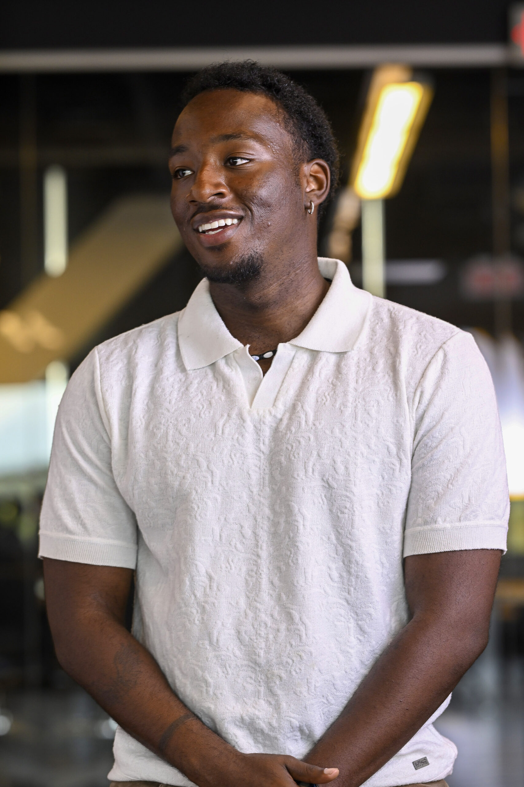 A man wearing a white short-sleeve polo shirt stands indoors, smiling with his hands clasped in front of him.