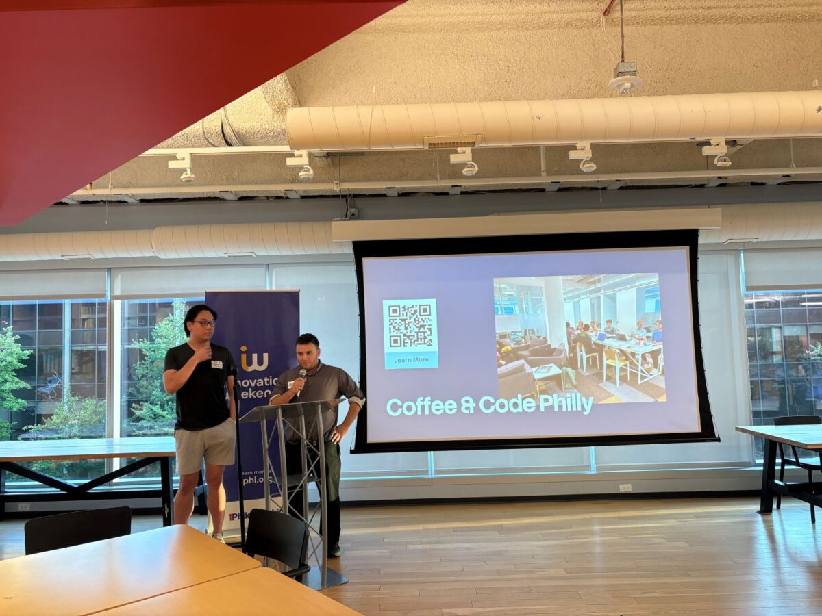 Two people stand at a podium presenting a slide titled “Coffee & Code Philly” with a QR code, in a modern room with large windows and exposed ceiling pipes.
