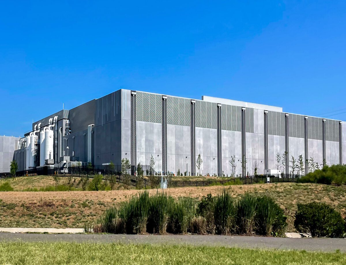 Large, modern data center building with cooling units on the side, surrounded by grass and landscaping under a clear blue sky.