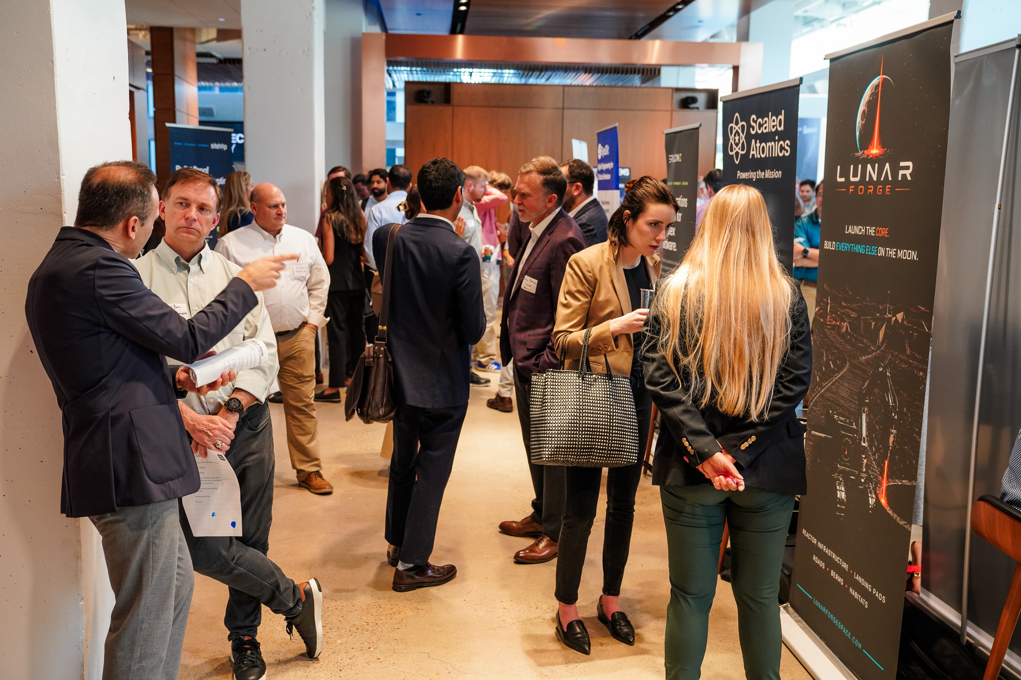People in business attire converse and network at a professional indoor event with informational booths and banners displayed along the sides of the room.