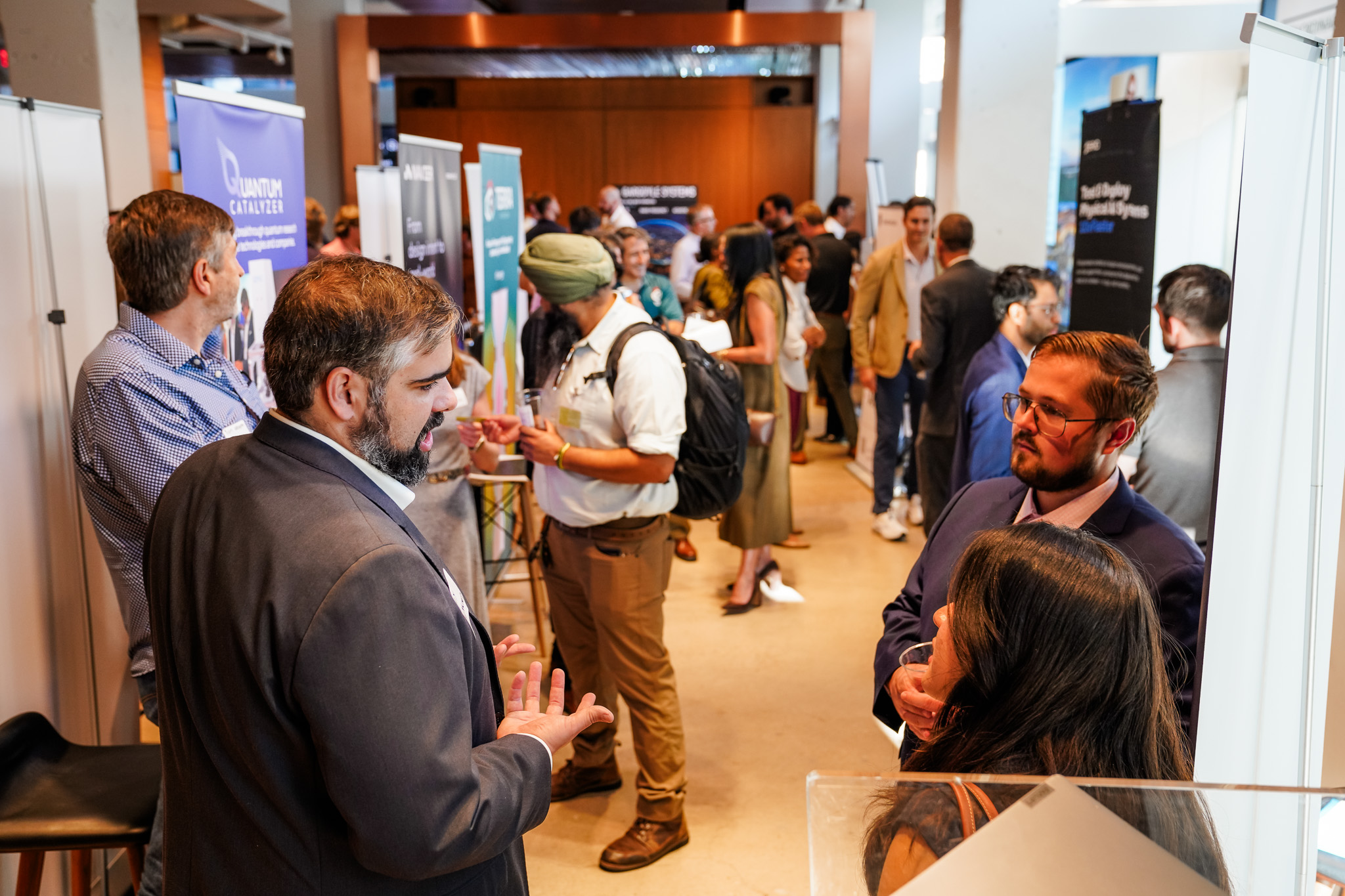 People networking at a professional event, standing in groups and talking near informational booths in a well-lit indoor space.
