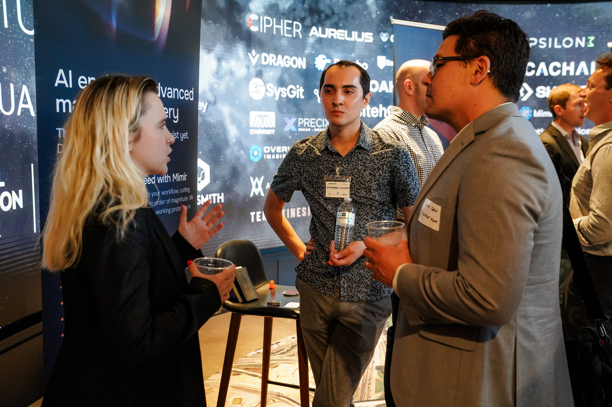 Three people stand and converse at a professional event, holding drinks, with a display of technology company logos in the background.