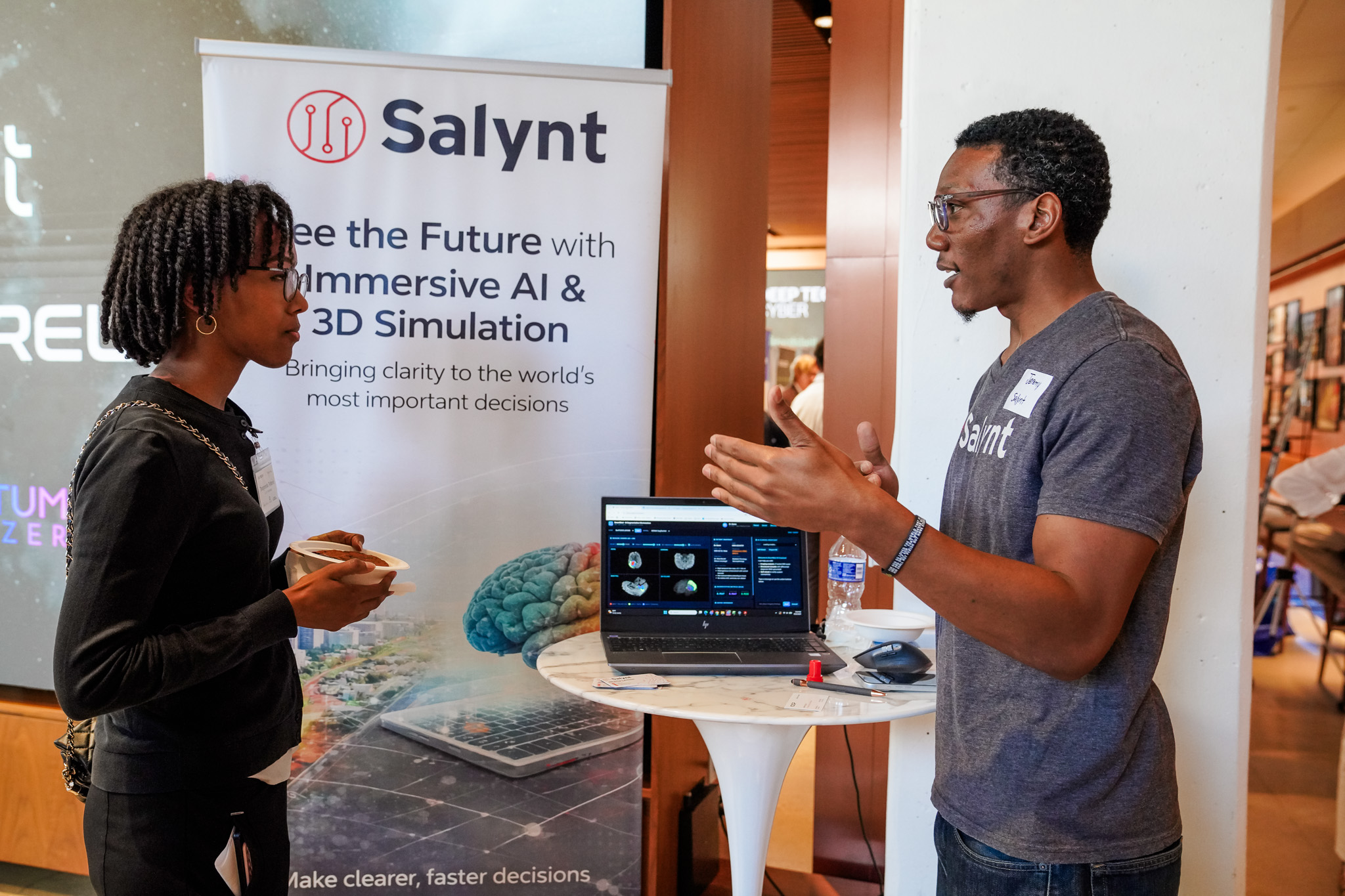 Two people talk at a booth with a Salynt banner and a laptop displaying AI and 3D simulation graphics. One person holds coffee while the other gestures during the conversation.
