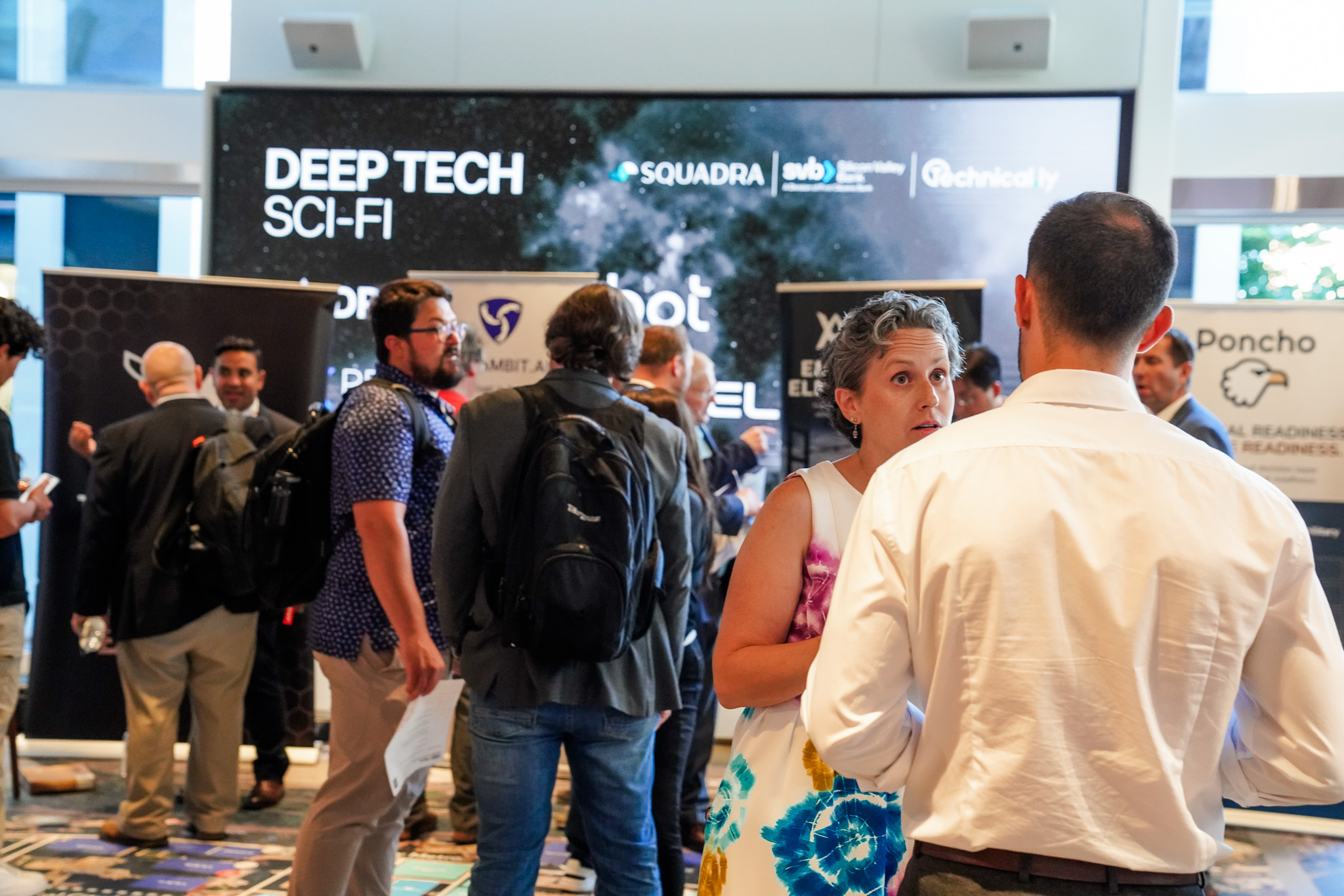 People converse at a tech conference with banners reading “Deep Tech Sci-Fi” in the background. Some attendees wear business attire, and others carry backpacks.