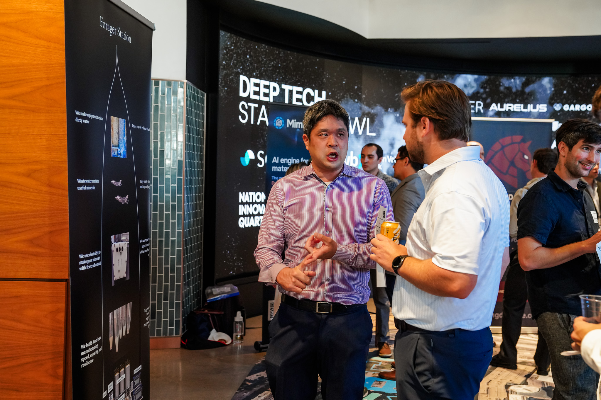 Two men talk at a tech event near a display board, while other attendees mingle in the background. A digital screen displays “DEEP TECH” and company logos.