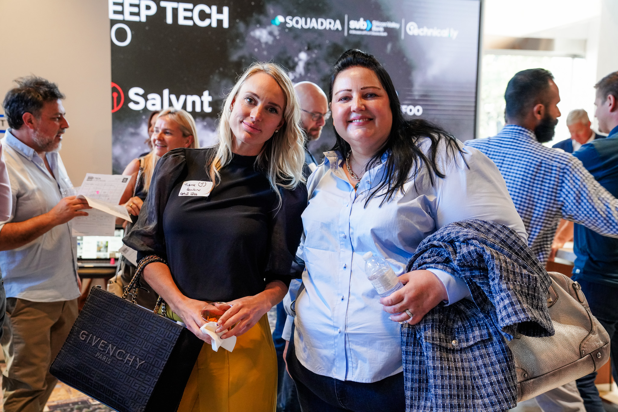 Two women pose and smile at a tech conference; people mingle in the background in front of a large event display screen.