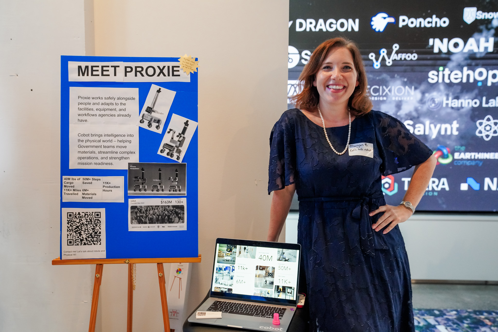 A woman stands next to a display board labeled “Meet Proxie” and a laptop, presenting information at an indoor event.
