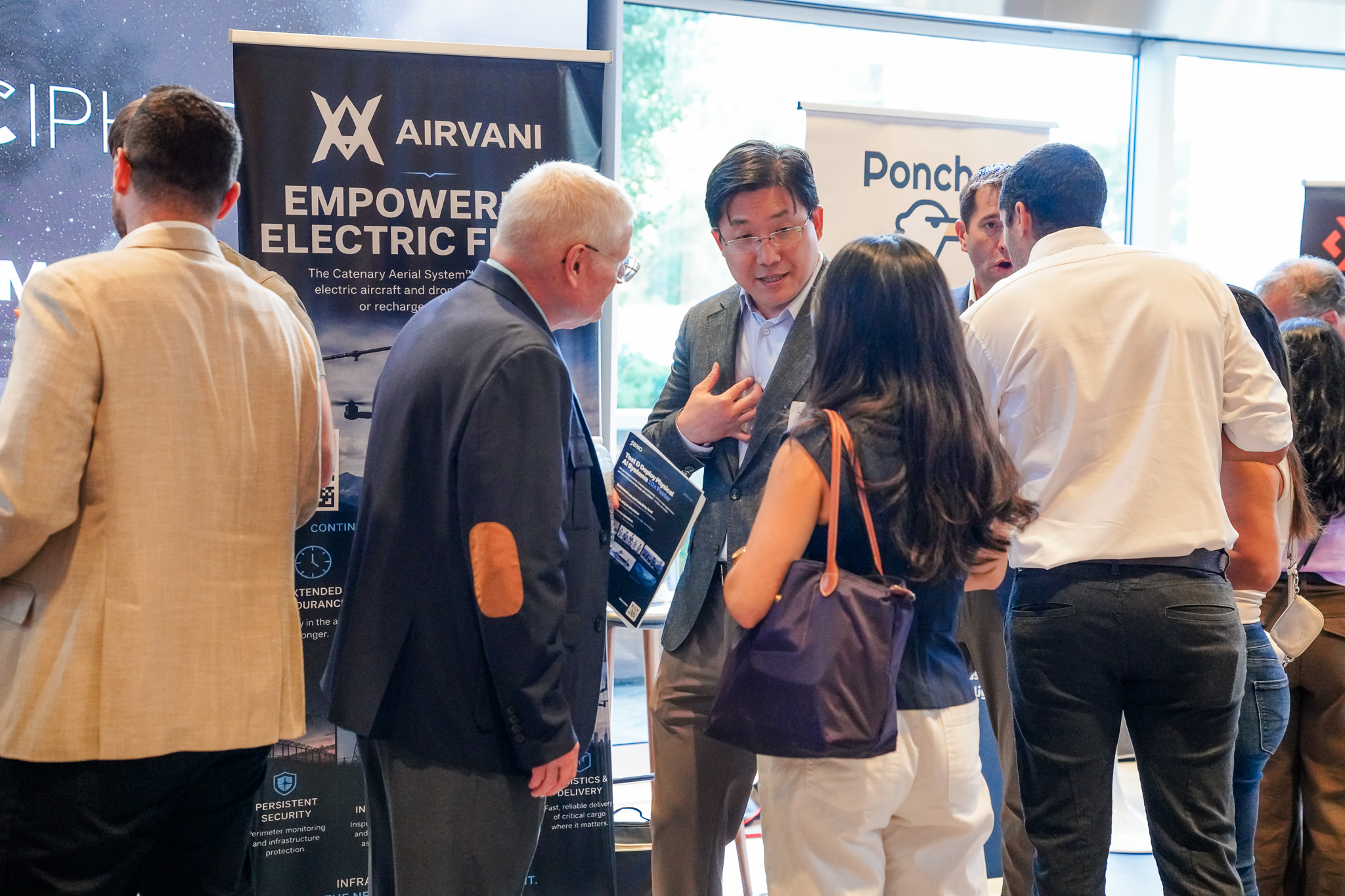 People stand and converse at an indoor business event, with informational banners about electric flight and security services visible in the background.