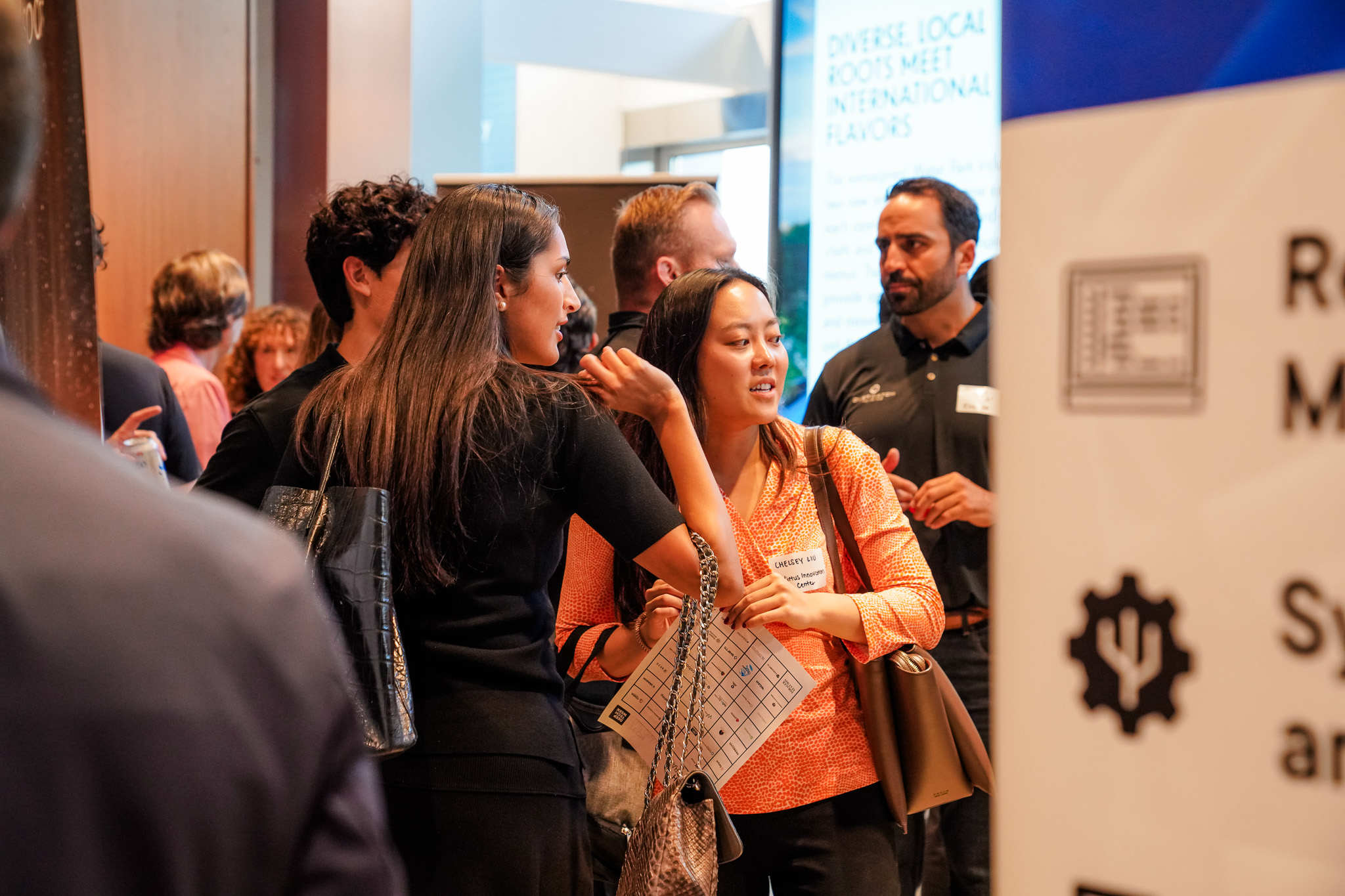 People gather at an indoor networking event, talking and holding papers; informational signs and banners are visible in the background.