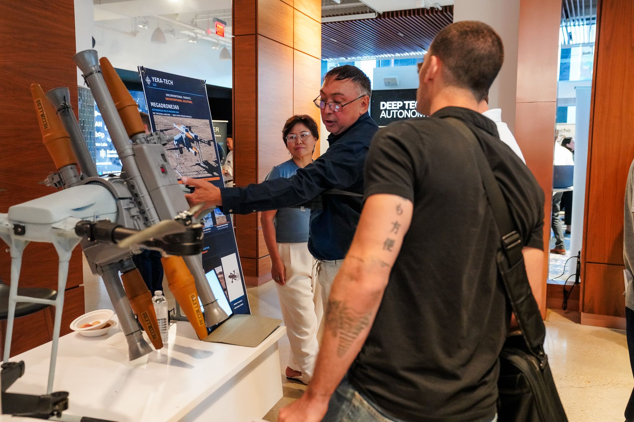 Three people examine a drone display at a technology exhibit. One person gestures toward the drone while informational posters are visible in the background.