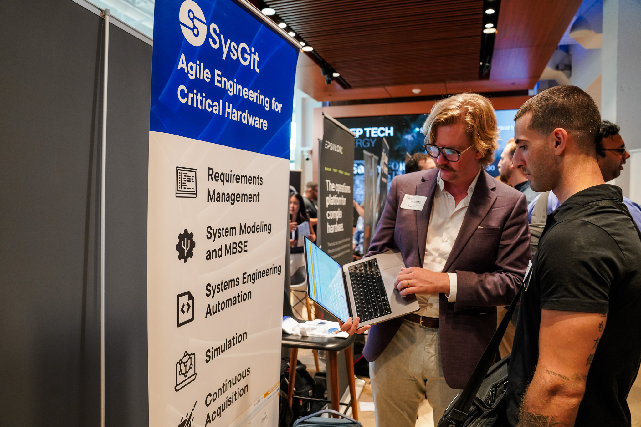 Two men stand by a SysGit booth at an event; one holds a laptop while discussing agile engineering services displayed on a large banner.
