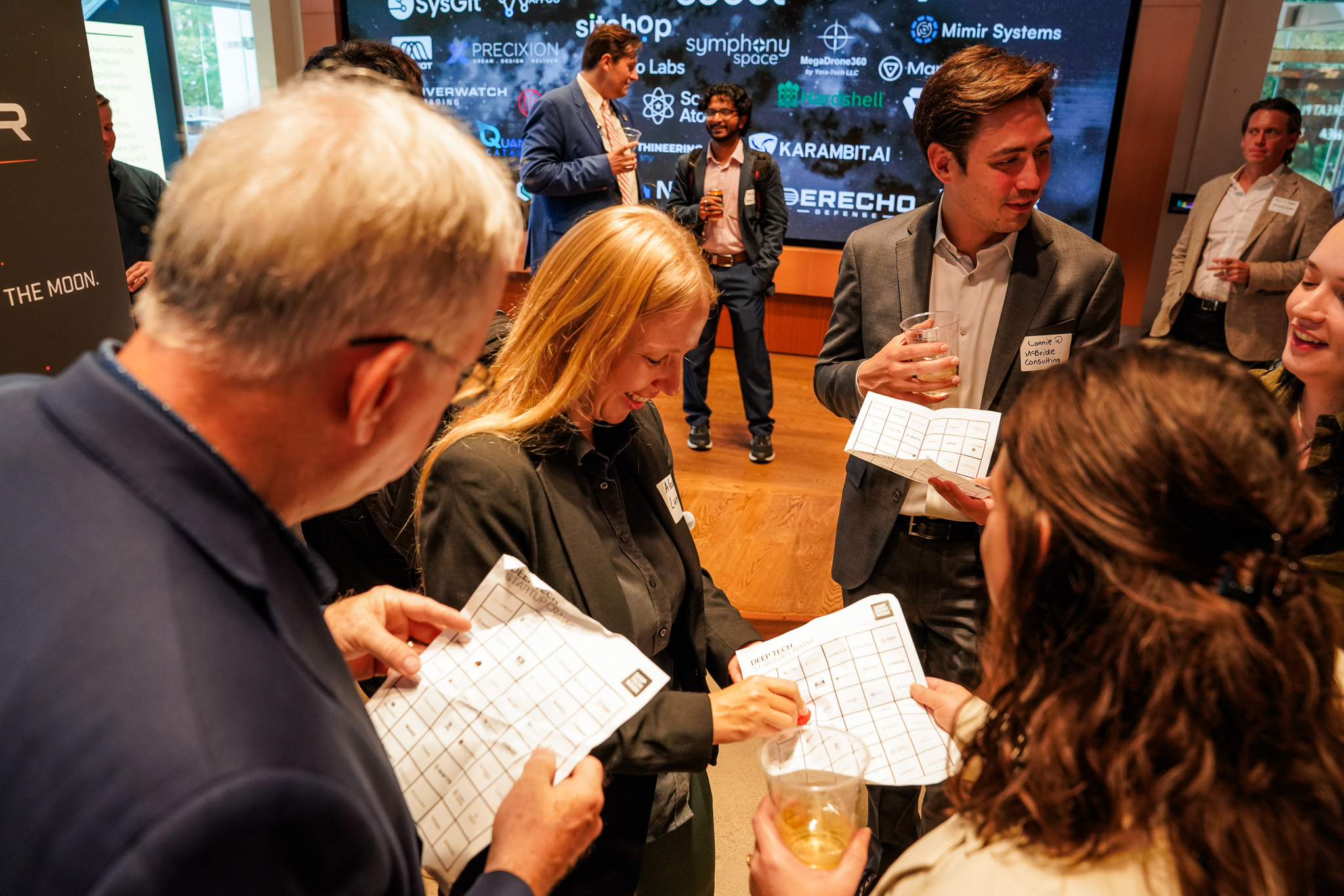 Several people stand together holding bingo cards and drinks at a networking event, with company logos displayed on a large screen in the background.