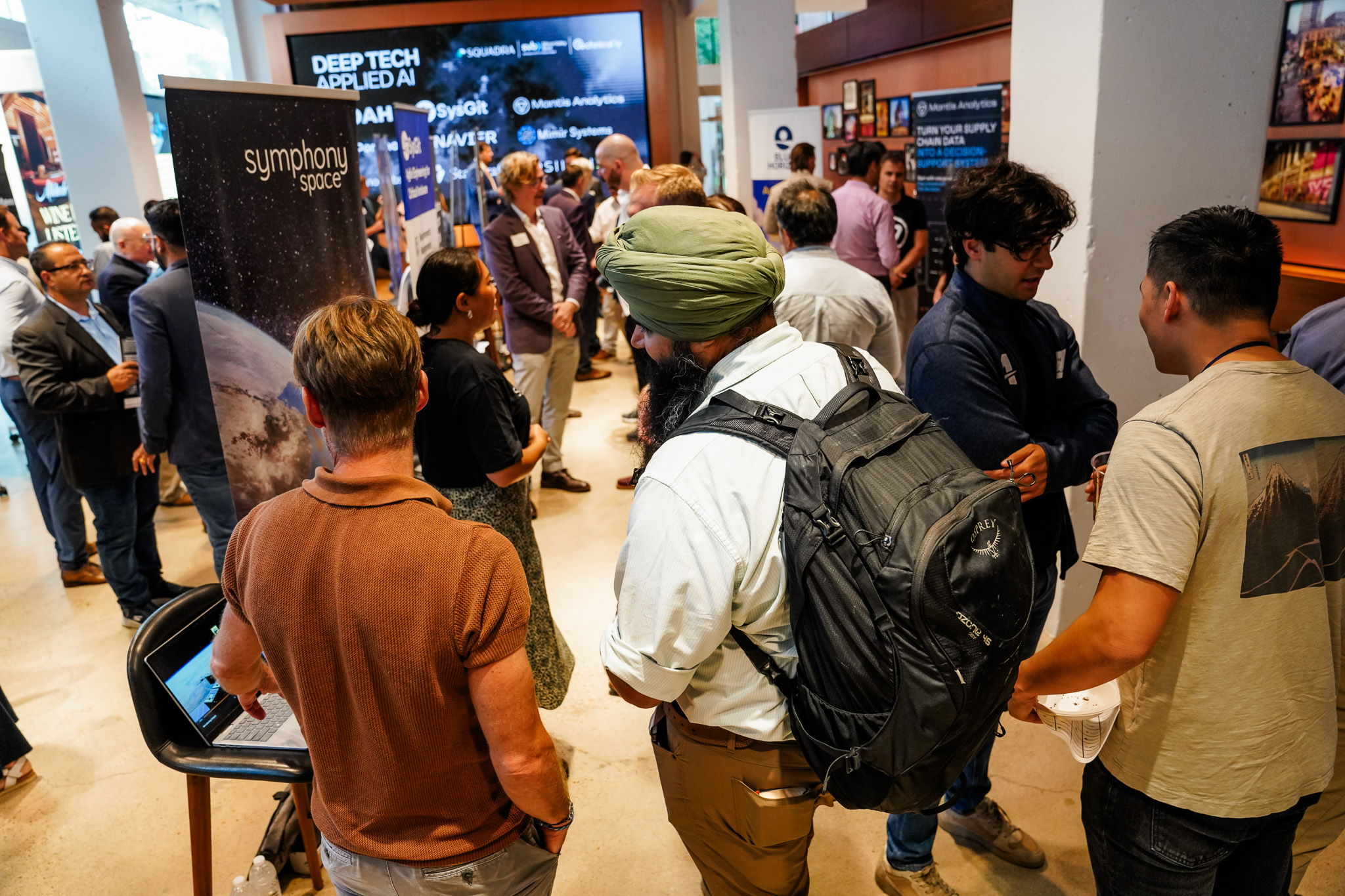 People gather and network at a tech event, standing near company booths with banners and informational displays in a modern indoor space.