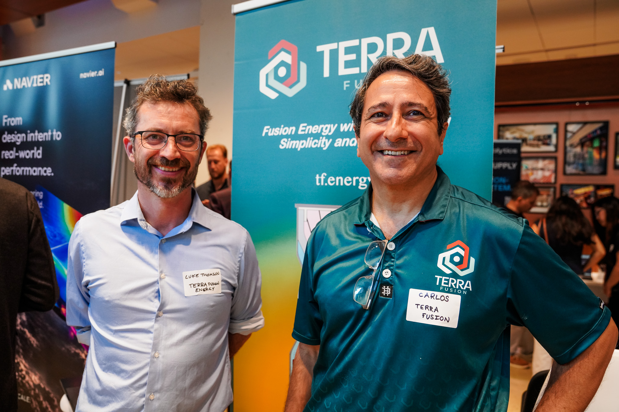 Two men stand smiling at an event, both wearing name tags, in front of a “Terra Fusion” promotional banner with informational booths in the background.
