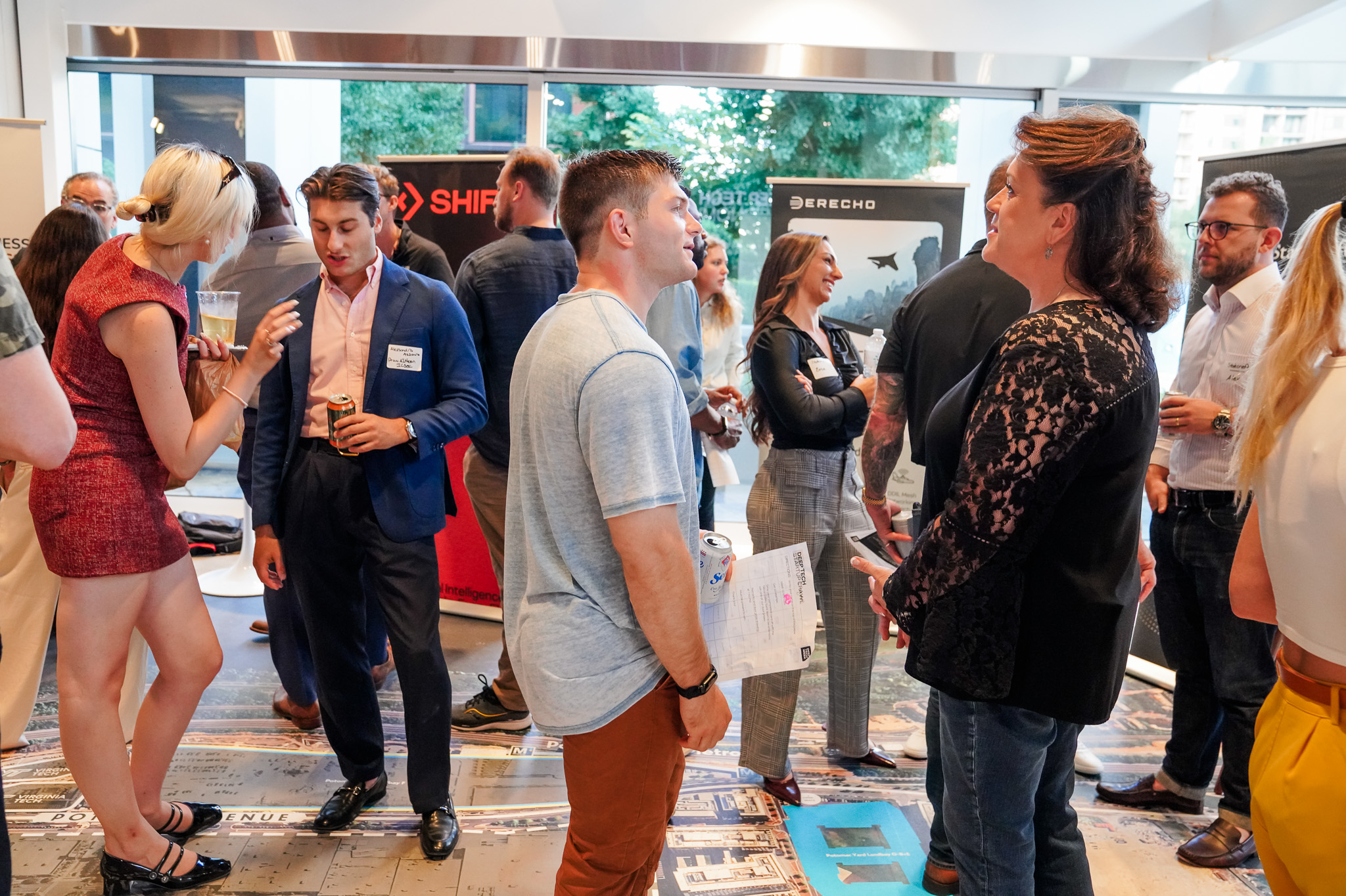 People stand and talk in small groups at an indoor networking event, holding papers and drinks with display banners and natural light from large windows in the background.