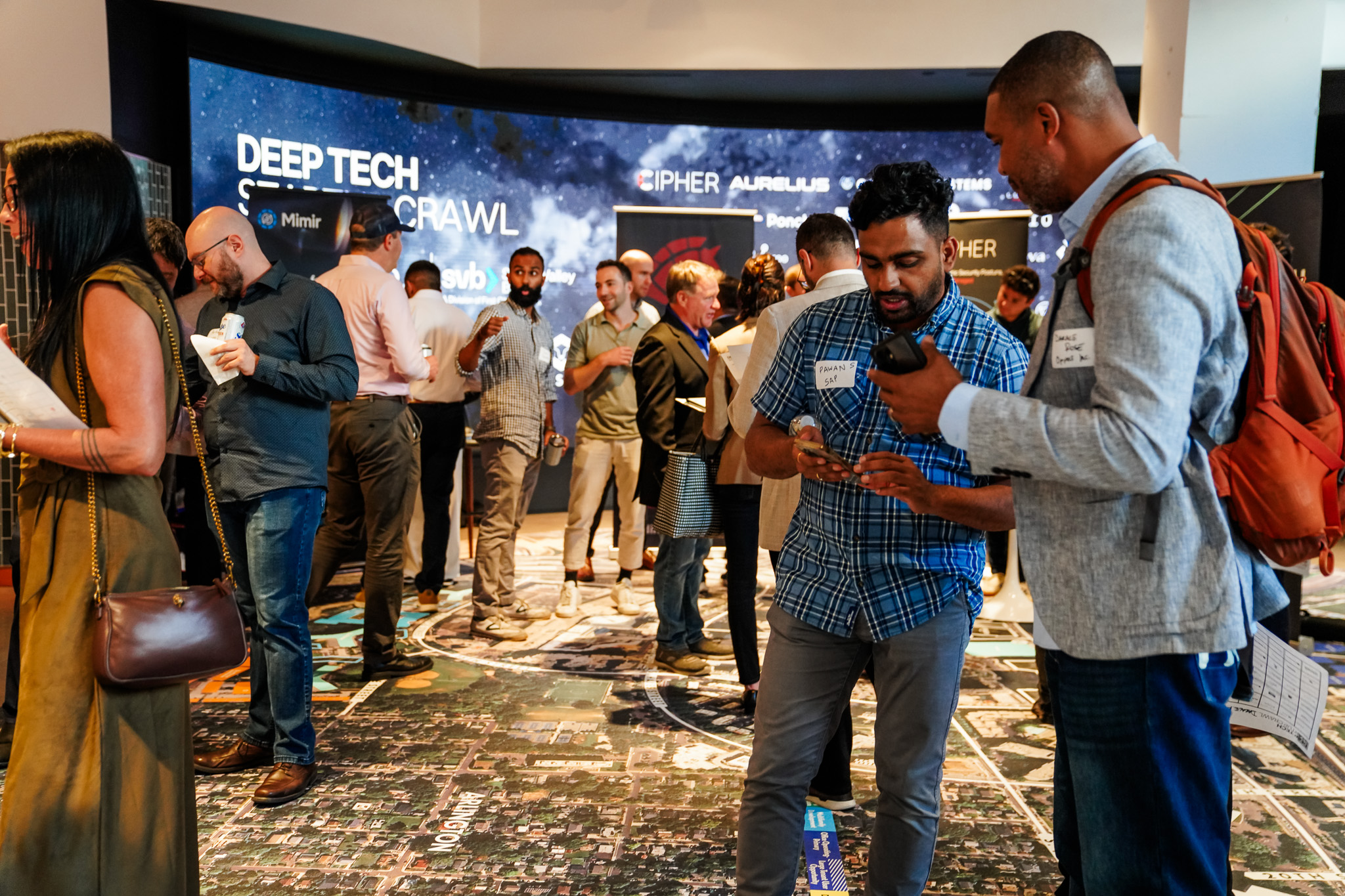 People network and socialize at a tech event, standing on a floor map, with event branding and company logos displayed on a screen in the background.