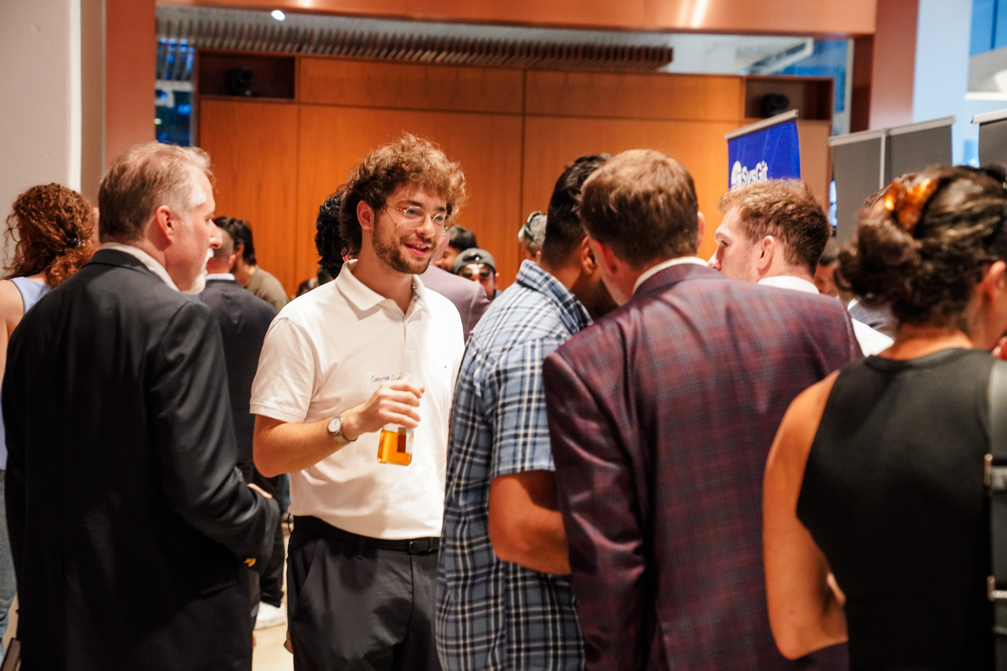 A group of people stand and talk at an indoor networking event; one person in a white shirt holds a drink and smiles while engaging in conversation.
