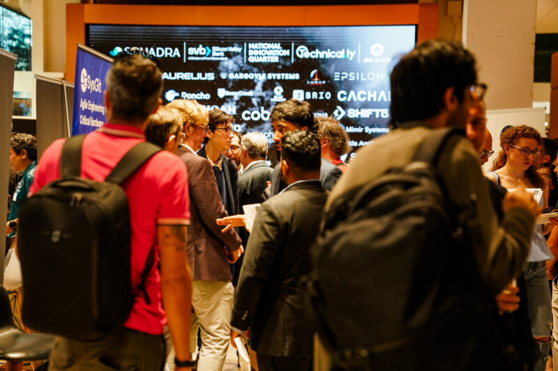 People gather and network at an indoor event with sponsor logos displayed on a screen in the background.