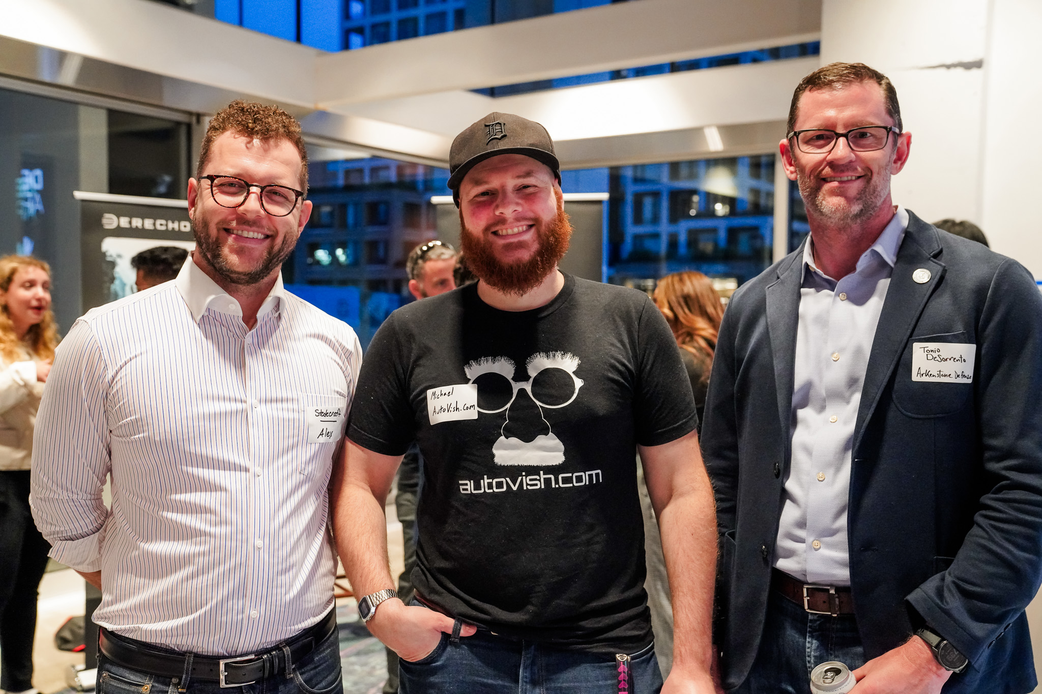 Three men stand indoors at an event, two wear glasses and button-up shirts, the one in the middle wears a black cap and T-shirt with white graphics. Other people mingle in the background.
