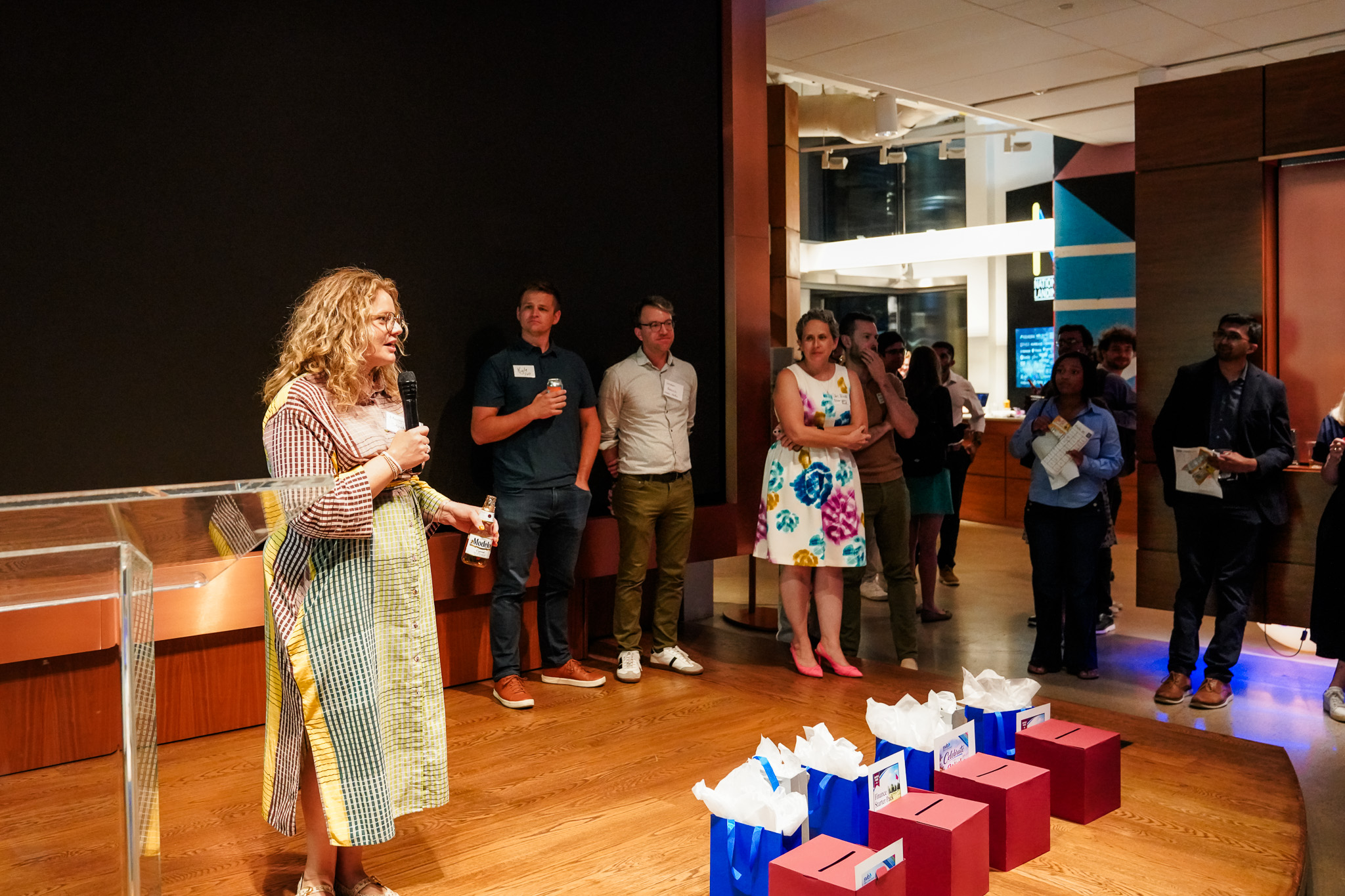 A woman with curly hair speaks at a podium in a modern office space, holding a microphone and a bottle. A group of people stand on a wood floor watching her, with gift bags in the foreground.