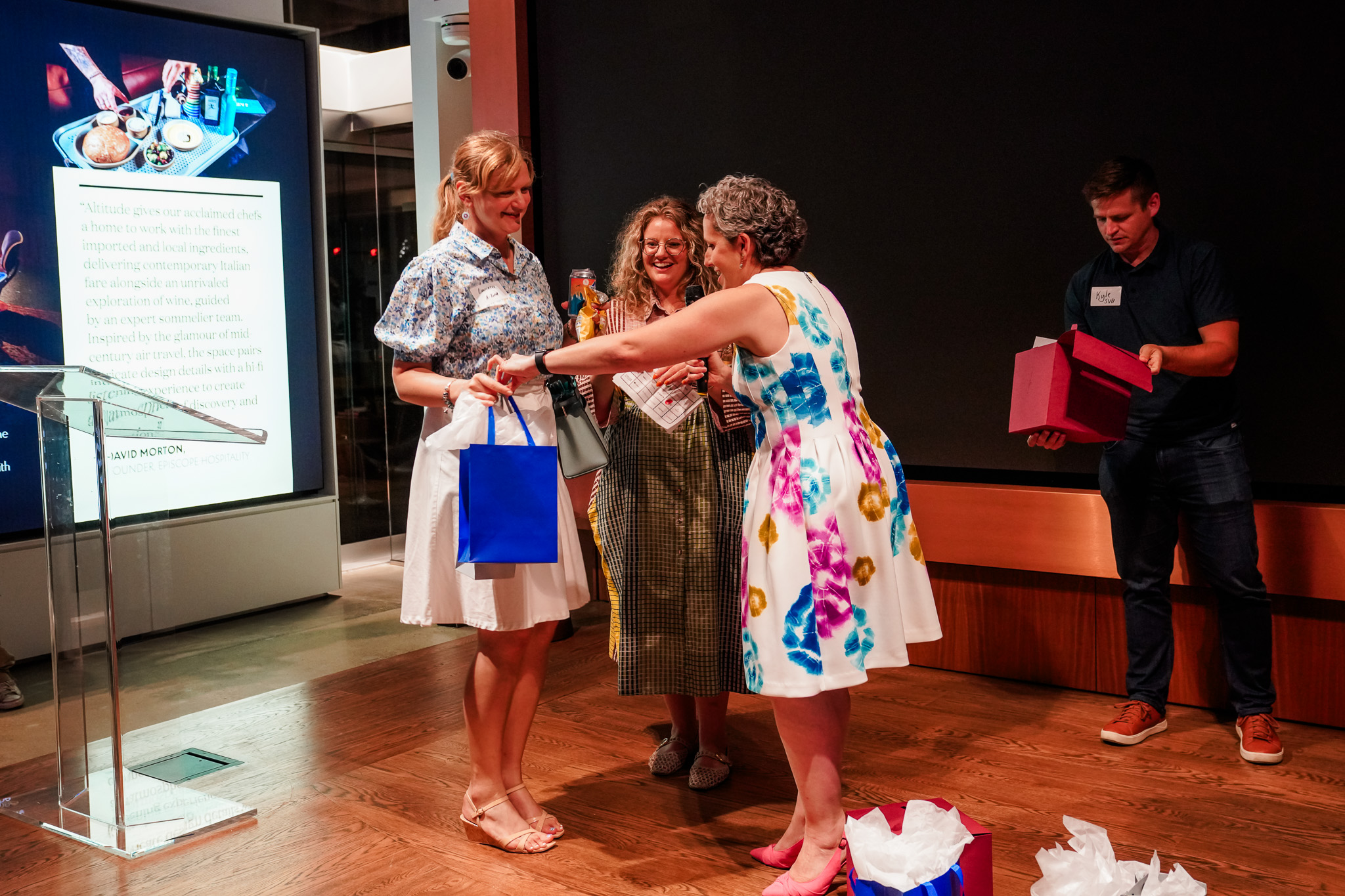 Four adults stand on a wooden floor exchanging gift bags and boxes; one woman hands a blue bag to another, while a man in the background holds a purple box.