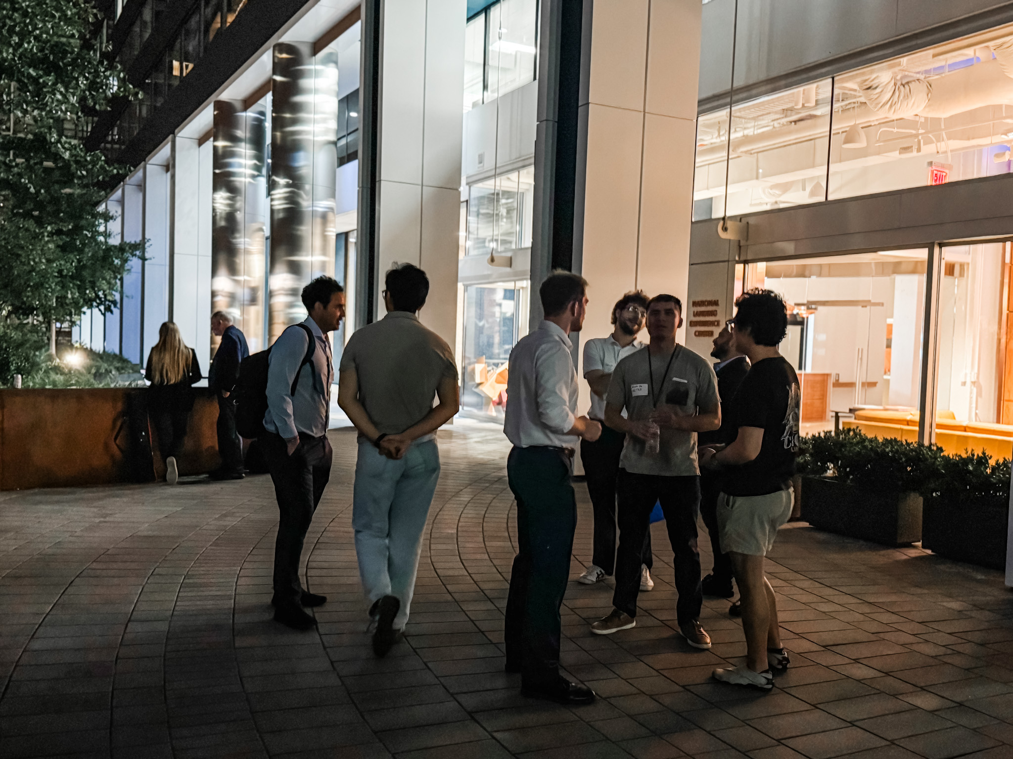 A group of people stand and talk outside a modern building at night, with lights illuminating the area and large windows visible behind them.