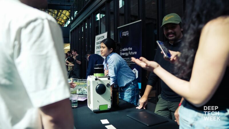 People stand around a table displaying a white device at an indoor event with “Deep Tech Week” branding visible in the corner.