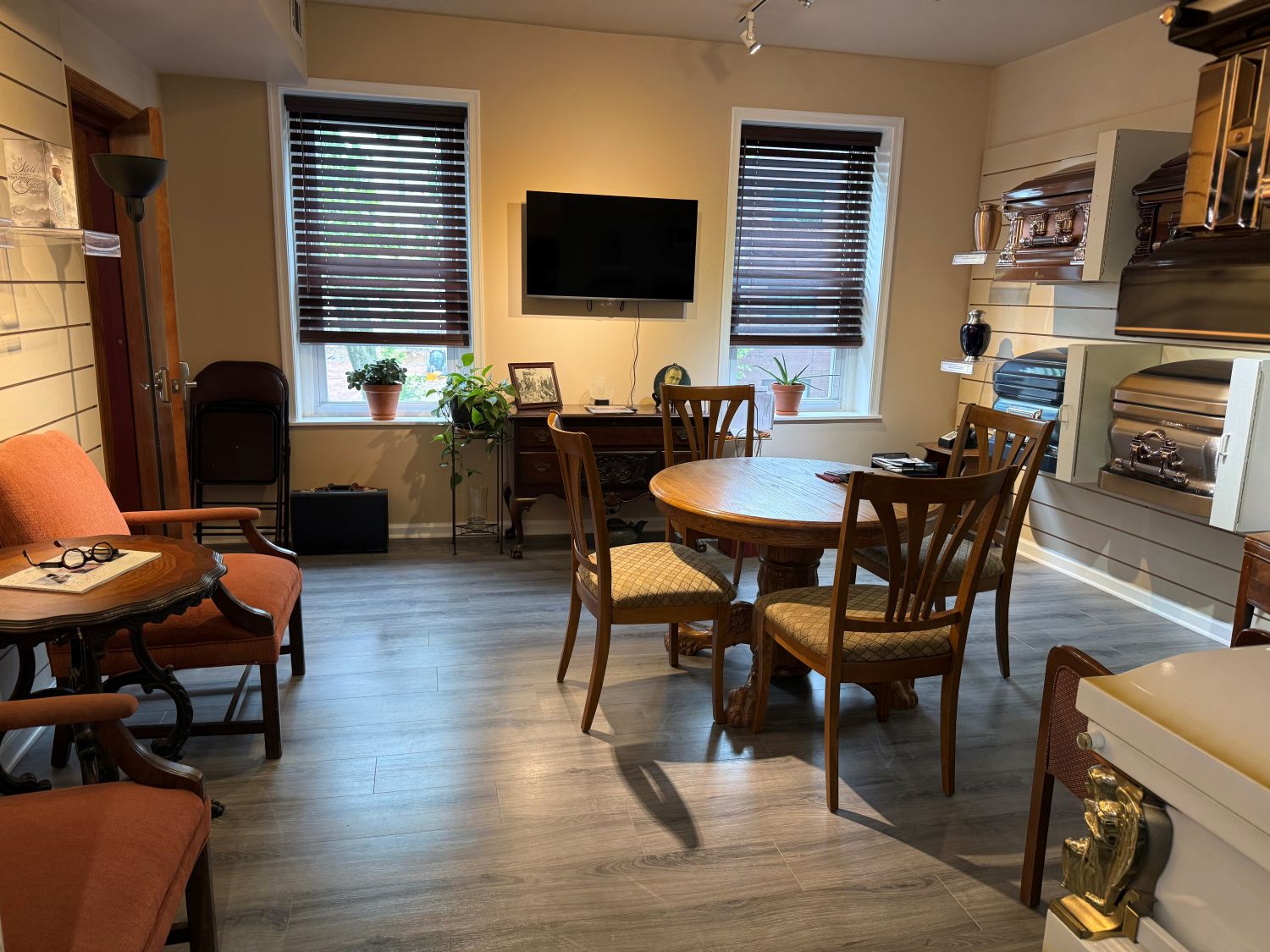 A small room with wooden chairs, two tables, a TV on the wall, plants by two windows, and urns and caskets displayed on shelves.