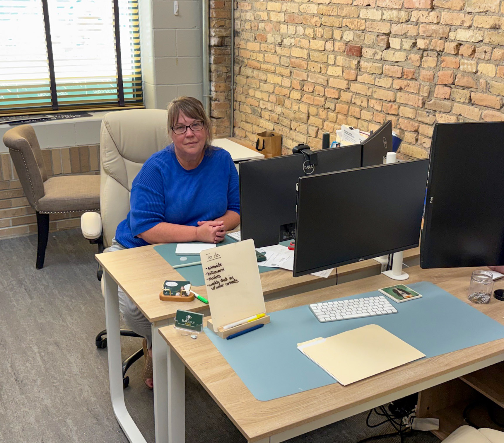 A person in a blue shirt sits at a desk with multiple monitors, papers, and office supplies in a workspace with brick walls and a window.