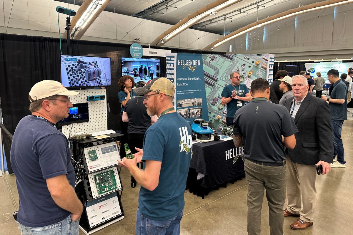 People converse at a technology trade show booth with monitors, circuit boards, and informational displays set up under a high ceiling.