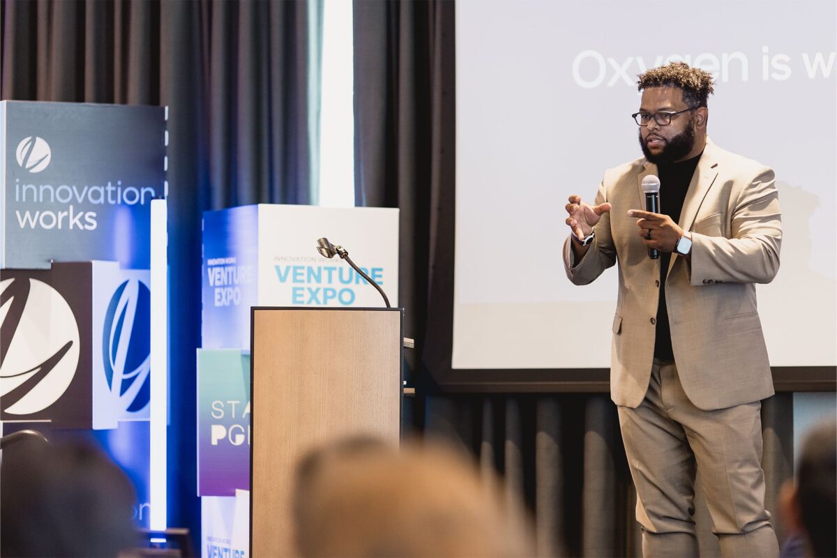 A man in a tan suit speaks on stage at a conference, standing beside a podium with “Venture Expo” banners in the background.