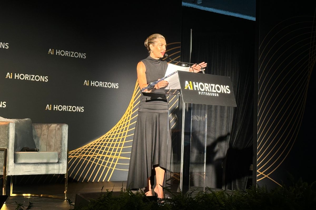 A woman stands at a podium labeled “AI HORIZONS Pittsburgh,” speaking at an event, with a black and gold backdrop behind her.