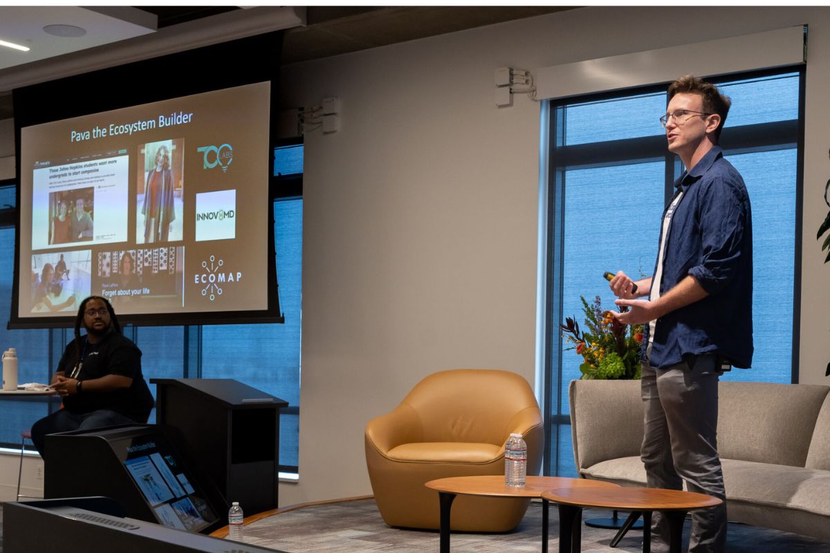 A person stands and presents slides about an ecosystem builder to an audience; another person sits at a table nearby in a modern conference room.
