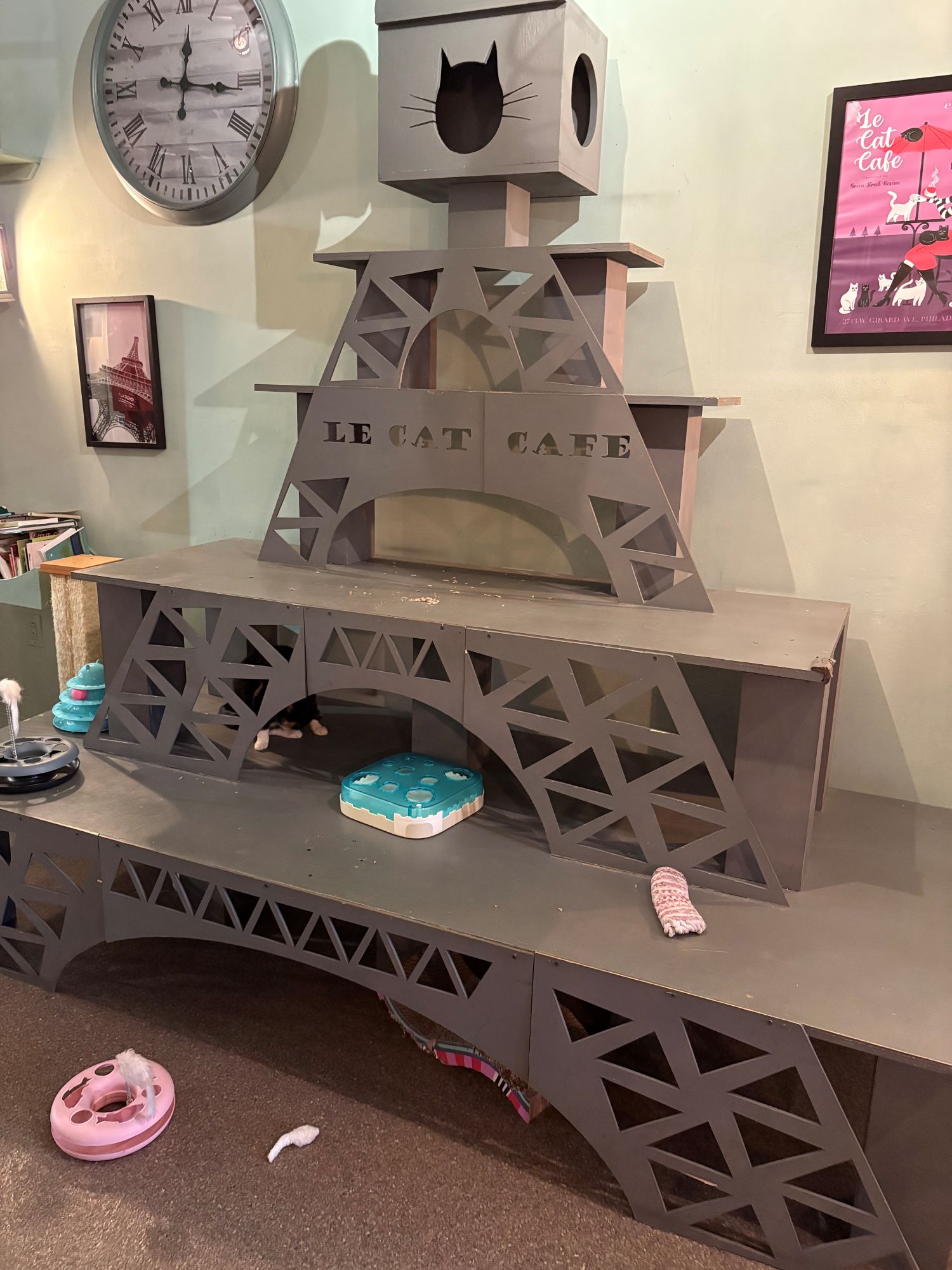 A cat play structure shaped like the Eiffel Tower with “LE CAT CAFE” written on it, various cat toys, and a wall clock and poster in the background.