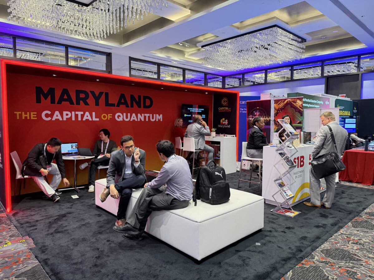 People sit and talk at the Maryland “Capital of Quantum” booth at a tech conference, surrounded by other exhibition booths and displays in a brightly lit hall.